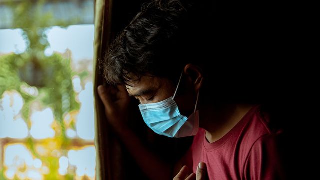 Man in a red shirt wearing a medical mask, coughing while standing near a window with sunlight streaming in 