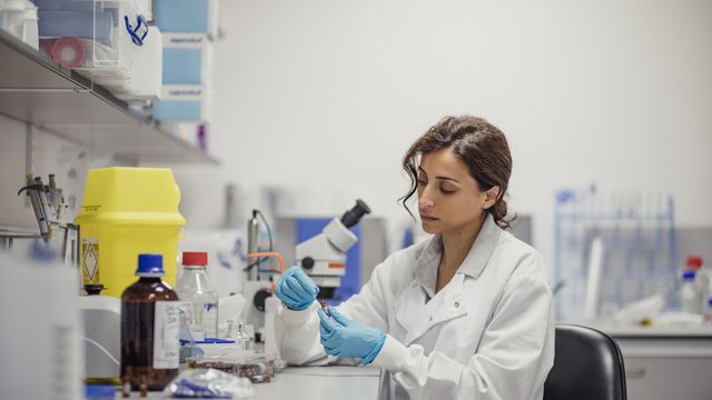 Female scientist in a laboratory wearing a white coat and blue gloves, carefully handling a vial with a microscope and lab equipment in the background