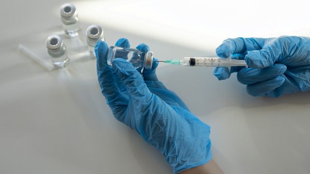 Gloved hands drawing liquid from a vial into a syringe, with additional vials in the background, symbolizing vaccine preparation