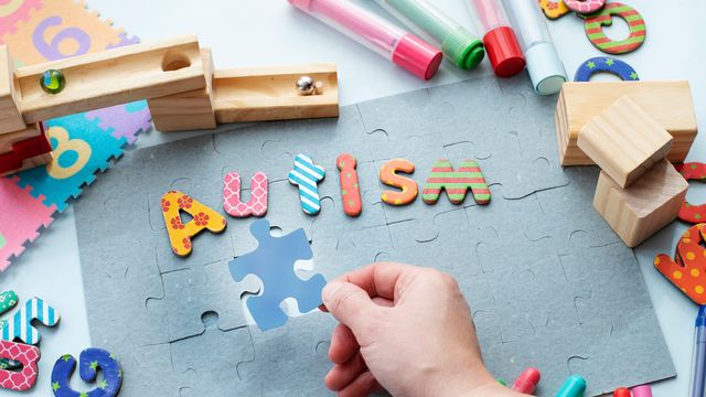 Child’s hand placing puzzle piece next to colorful letters spelling "autism" amid educational toys.