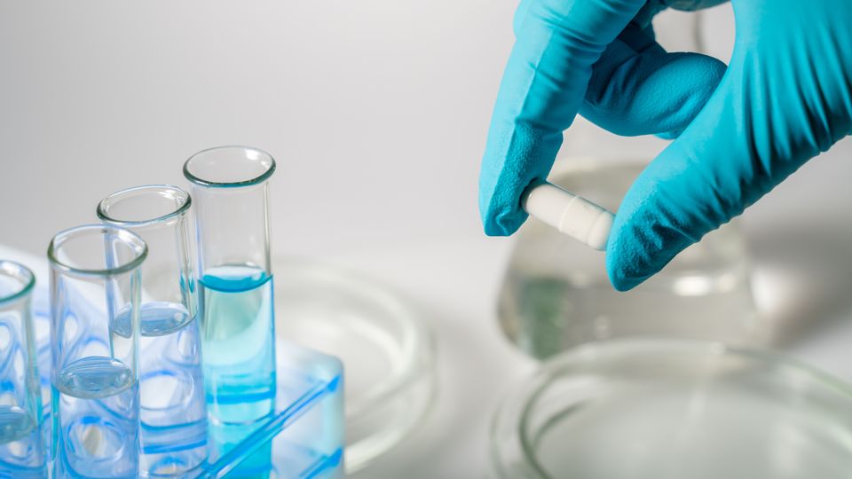 A gloved hand holds a white capsule between thumb and finger. Test tubes and petri dishes visible.