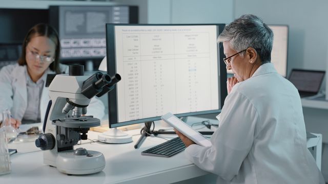 Two scientists in a laboratory analyzing data on a computer and tablet beside a microscope.
