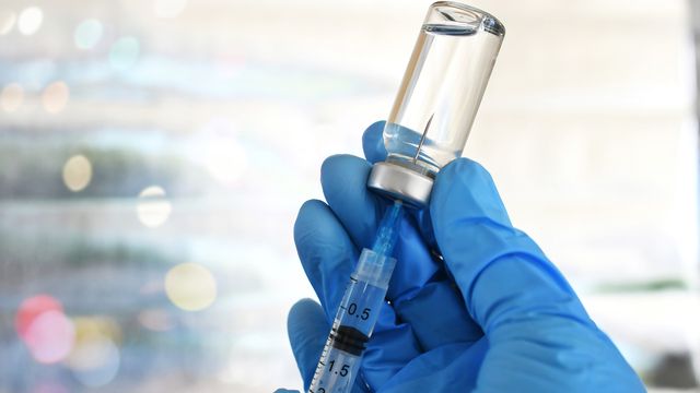 Gloved hand drawing liquid from a vial with a syringe, representing advancements in synthetic biology.