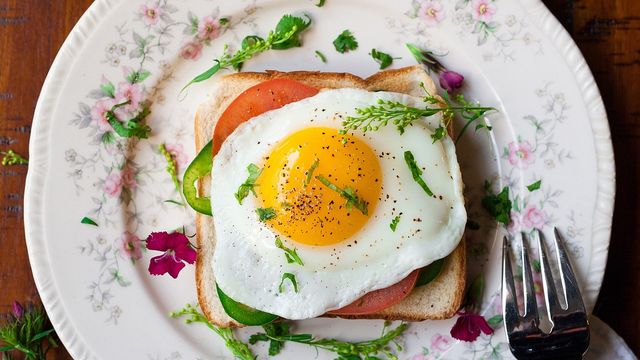 Overhead shot of a fried egg on tomato and toast with herbs and seasoning laying on a floral plate with a fork at the edge.