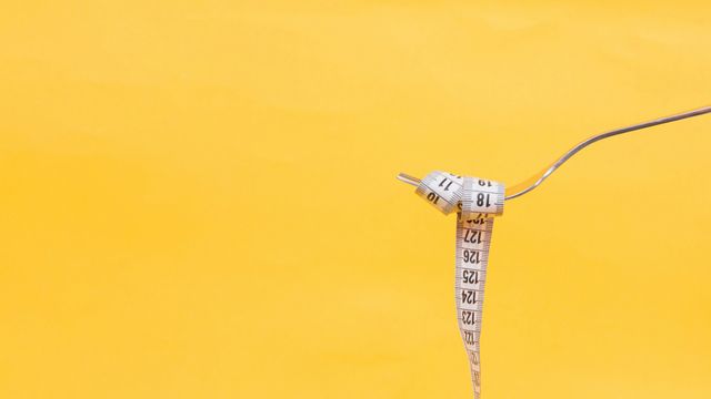 Fork holding a curled measuring tape against a yellow background symbolizing weight loss.