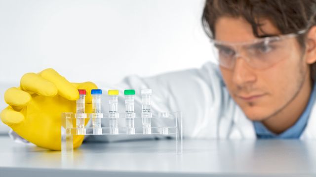 Scientist wearing safety goggles and a yellow glove handling labeled vials in a laboratory