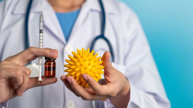 A doctor in a lab coat holding a vial of medicine, a needle and a model of a virus.