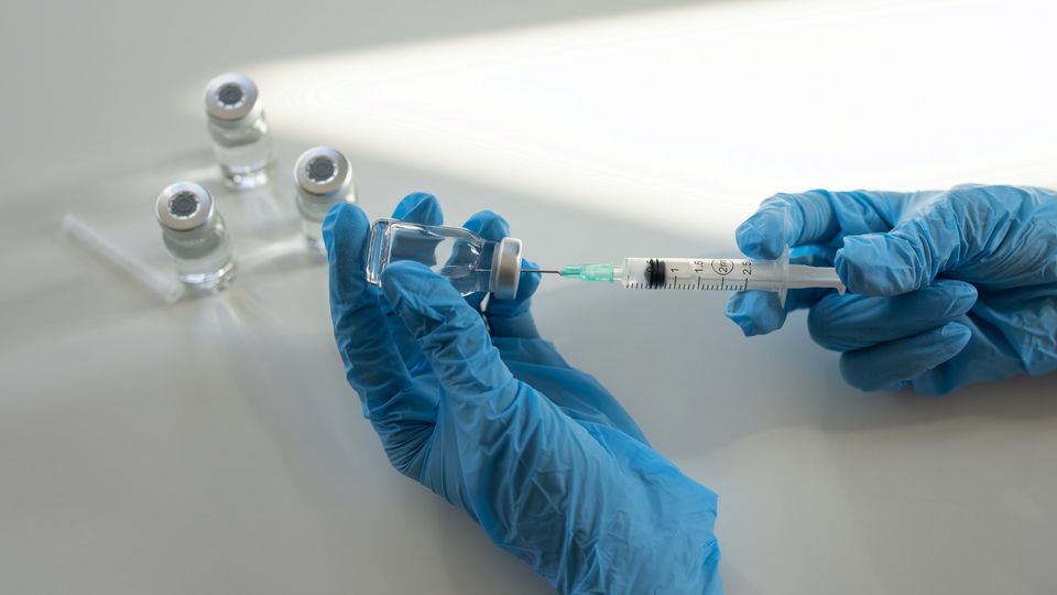 Two gloved hands hold a syringe and a vial, the former piercing the other. It seems a vaccination is being drawn up.