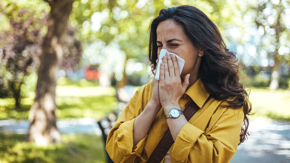 Woman sneezing into a tissue outdoors, showing symptoms of hay fever during allergy season.
