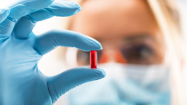 A female scientist in a mask and blue gloves holding up a red drug pill, representing drug discovery.