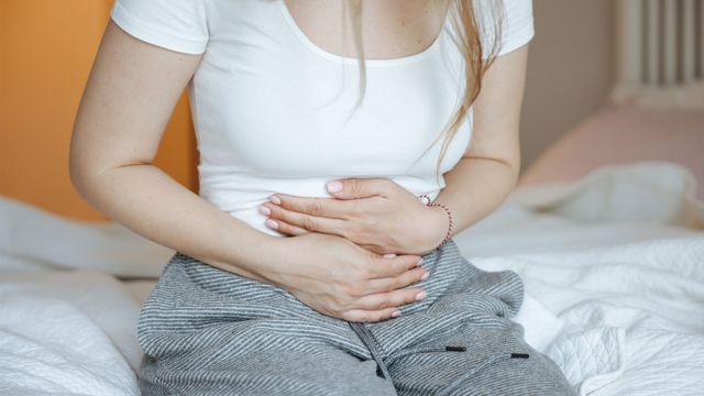 Woman sat on edge of bed holding stomach in pain. 