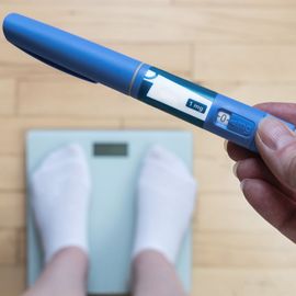 Hand holding a weight loss drug injector above a bathroom scale with feet in socks. 