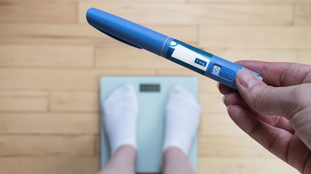 Hand holding a weight loss drug injector above a bathroom scale with feet in socks. 