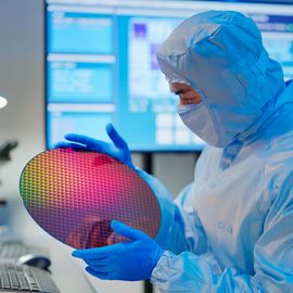Scientist in a coverall, mask and gloves holding a wafer in a semiconductor manufacturing facility and examining it. 