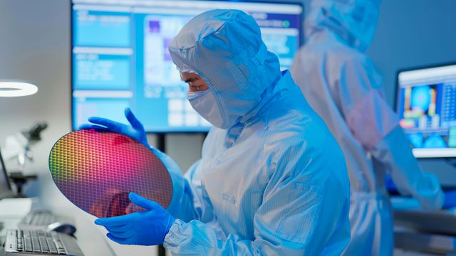Scientist in a coverall, mask and gloves holding a wafer in a semiconductor manufacturing facility and examining it. 