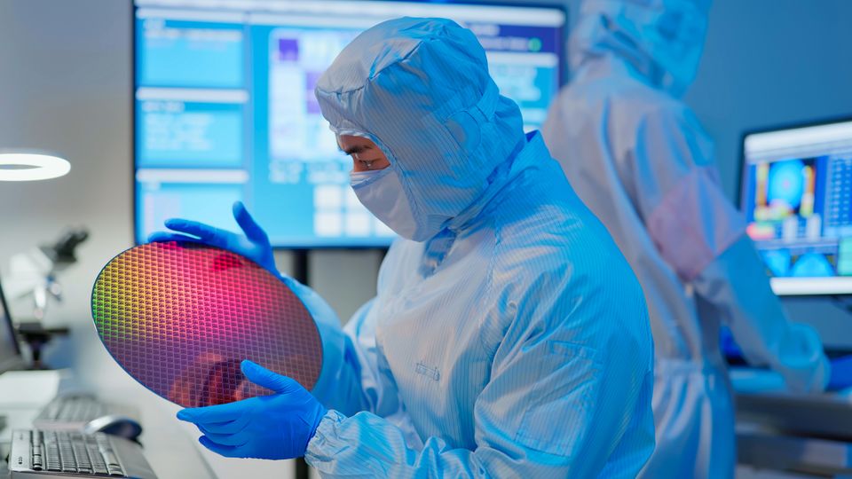 Scientist in a coverall, mask and gloves holding a wafer in a semiconductor manufacturing facility and examining it.